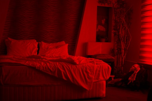 A bedroom with a disheveled bed that has two pillows and white sheets. There is a wavy pattern on the back wall behind the bed. To the right, there is a plant next to a small table. On the wall above the table is an art piece depicting a person. The room is dominated by a red lighting, casting a tint over everything.