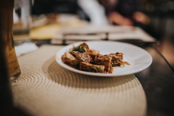 A white plate rests on a woven placemat with pieces of food that appear to be meat with sauce and green garnish. The background is blurred, providing a warm and inviting atmosphere.