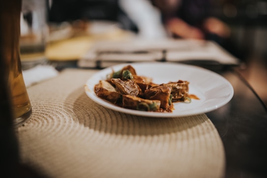 A white plate rests on a woven placemat with pieces of food that appear to be meat with sauce and green garnish. The background is blurred, providing a warm and inviting atmosphere.