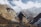A hiker stands on a mountainous terrain holding a walking stick, gazing at the vast, snow-capped mountains in the background. The scene includes rugged cliffs, sparse vegetation, and a few small, blue-roofed structures in a valley below.
