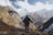 A hiker stands on a mountainous terrain holding a walking stick, gazing at the vast, snow-capped mountains in the background. The scene includes rugged cliffs, sparse vegetation, and a few small, blue-roofed structures in a valley below.