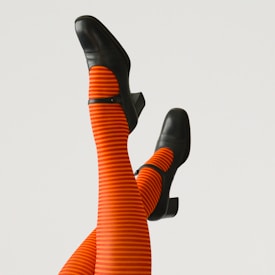 Black heeled shoes are paired with bright orange and red striped tights, with legs raised against a neutral background.