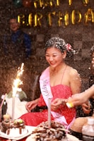 A person wearing a red dress and a 'Happy Birthday' sash is smiling amidst a festive atmosphere. There is a birthday cake with lit sparklers in front of them, and confetti or snow-like substance is falling around. Other people are partially visible in the background, and golden 'Happy Birthday' balloons are displayed.
