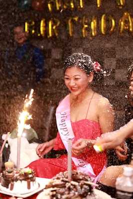 A person wearing a red dress and a 'Happy Birthday' sash is smiling amidst a festive atmosphere. There is a birthday cake with lit sparklers in front of them, and confetti or snow-like substance is falling around. Other people are partially visible in the background, and golden 'Happy Birthday' balloons are displayed.