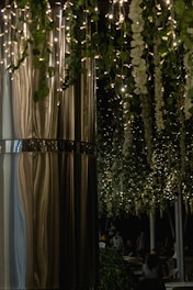 A dimly lit indoor or outdoor space showcasing elegant hanging decorations. Twinkling fairy lights interlace with lush green foliage and cascading white flowers. A smooth, reflective metallic surface forms a large column on the left, adding a modern touch. Several people can be seen sitting quietly in the background, partially obscured by shadows.