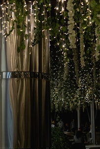 A dimly lit indoor or outdoor space showcasing elegant hanging decorations. Twinkling fairy lights interlace with lush green foliage and cascading white flowers. A smooth, reflective metallic surface forms a large column on the left, adding a modern touch. Several people can be seen sitting quietly in the background, partially obscured by shadows.