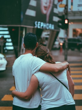 A couple stands close together, embracing, as they face a busy street with a large advertisement in the background featuring a model's portrait. It's a slightly overcast day, and the people appear to be relaxed and affectionate.