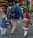 A family of four, including two adults and two children, walks together outside on a city street. The adults are wearing backpacks, and one child holds the hand of an adult. Brightly colored flowers are displayed in a store window in the background, suggesting a florist shop.