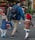 A family of four, including two adults and two children, walks together outside on a city street. The adults are wearing backpacks, and one child holds the hand of an adult. Brightly colored flowers are displayed in a store window in the background, suggesting a florist shop.