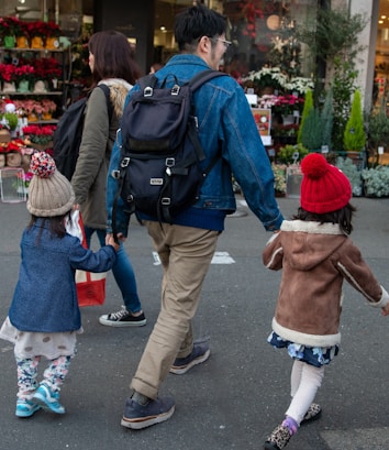 A family of four, including two adults and two children, walks together outside on a city street. The adults are wearing backpacks, and one child holds the hand of an adult. Brightly colored flowers are displayed in a store window in the background, suggesting a florist shop.