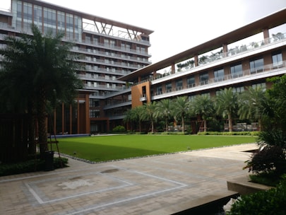 A modern building with multiple floors featuring large windows and a sleek design. There is a landscaped courtyard with well-maintained grass, palm trees, and other greenery. The architecture includes a mix of glass and wood elements, with outdoor balconies on some of the levels.