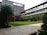 A modern building with multiple floors featuring large windows and a sleek design. There is a landscaped courtyard with well-maintained grass, palm trees, and other greenery. The architecture includes a mix of glass and wood elements, with outdoor balconies on some of the levels.