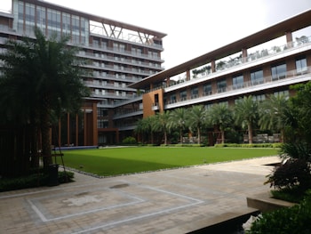 A modern building with multiple floors featuring large windows and a sleek design. There is a landscaped courtyard with well-maintained grass, palm trees, and other greenery. The architecture includes a mix of glass and wood elements, with outdoor balconies on some of the levels.