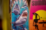 Two surfboards are depicted, one with an illustration of sharks, featuring a large shark with an open mouth and other smaller sharks around it. The other surfboard has a vibrant design with a silhouette of a surfer against a yellow and pink sunset background.