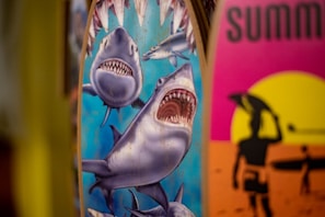 Two surfboards are depicted, one with an illustration of sharks, featuring a large shark with an open mouth and other smaller sharks around it. The other surfboard has a vibrant design with a silhouette of a surfer against a yellow and pink sunset background.