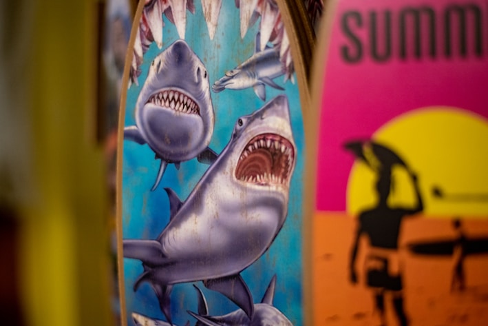 Two surfboards are depicted, one with an illustration of sharks, featuring a large shark with an open mouth and other smaller sharks around it. The other surfboard has a vibrant design with a silhouette of a surfer against a yellow and pink sunset background.