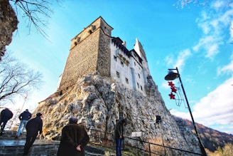 A large, historic castle is perched on a rocky hill under a bright blue sky. Several tourists are walking up a stone path towards the castle. Bare trees frame the scene, and a streetlamp adorned with red decorations stands in the foreground.