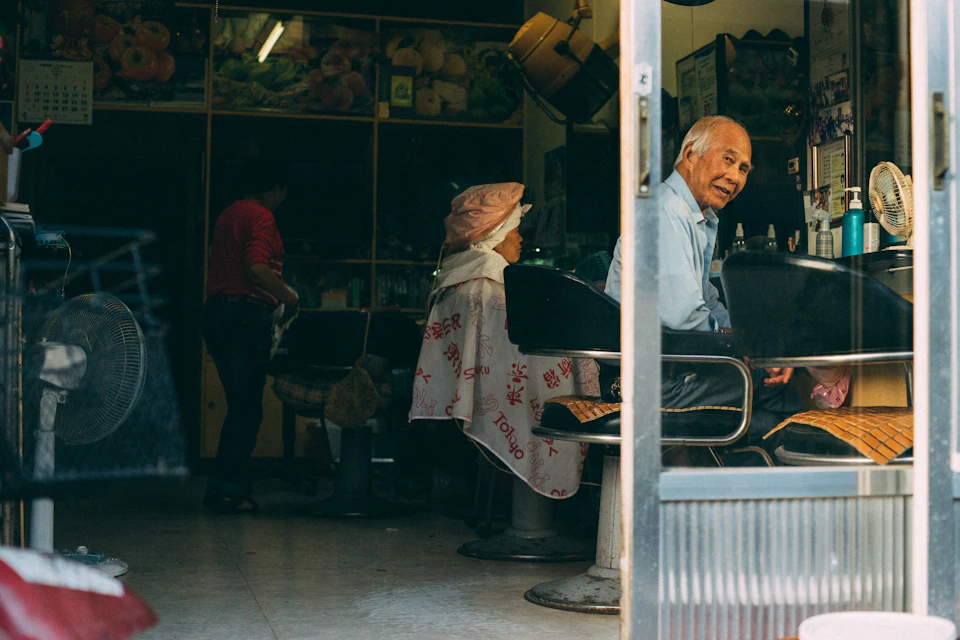 The image depicts a hair salon with three people inside, two seated and one standing behind a counter. The person on the left is wearing a red shirt and holding a bag, while the individual in the center has a pink hat on their head. On the right side of the photo, there's another person who appears to be a customer or an employee at the salon. The salon features various hair products displayed behind the counter, including bottles of shampoo and conditioner.
