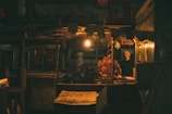 A dimly lit market stall with two people conversing under a single light bulb. The scene appears to be set indoors, with the focus on a man wearing a black shirt facing another person in a floral outfit. Several details such as hanging decorations and a van visible in the background add to the ambiance.