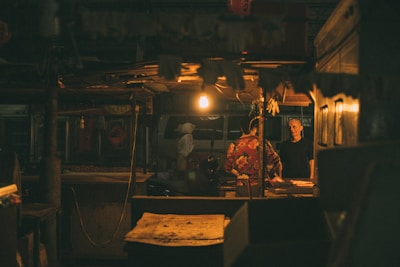 A dimly lit market stall with two people conversing under a single light bulb. The scene appears to be set indoors, with the focus on a man wearing a black shirt facing another person in a floral outfit. Several details such as hanging decorations and a van visible in the background add to the ambiance.