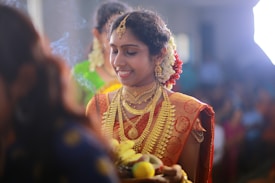 A woman adorned in traditional attire featuring elaborate gold jewelry stands, holding a tray with fruits. Her hair is decorated with jasmine flowers, and she wears a vibrant orange saree with intricate patterns. The background is slightly blurred, filled with soft lighting, suggesting a festive or ceremonial event.