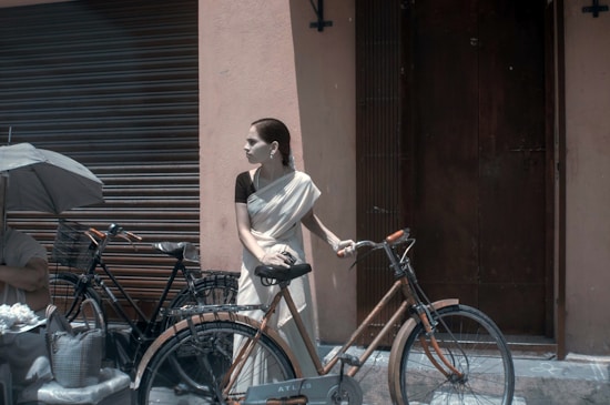 A woman in a white saree with a black blouse is standing next to a bicycle in an urban setting. She is looking to her right, with her left hand resting on the bicycle's handlebar. Behind her is a closed shutter and a dark brown door. To the left, a person is partially visible, holding an umbrella and sitting next to another bicycle.