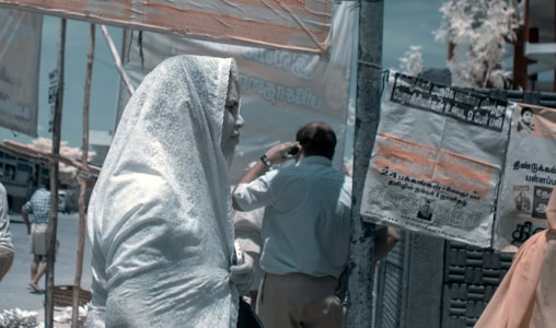 A person is standing on a busy street, draped in a light-colored fabric that covers their head and body. Several people are seen in the background, occupying themselves with different activities. Various posters and banners with Tamil script are visible on the walls and poles around, indicating a public place in a Tamil-speaking region.