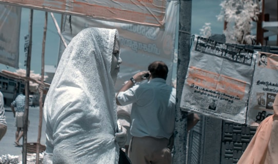 A person is standing on a busy street, draped in a light-colored fabric that covers their head and body. Several people are seen in the background, occupying themselves with different activities. Various posters and banners with Tamil script are visible on the walls and poles around, indicating a public place in a Tamil-speaking region.