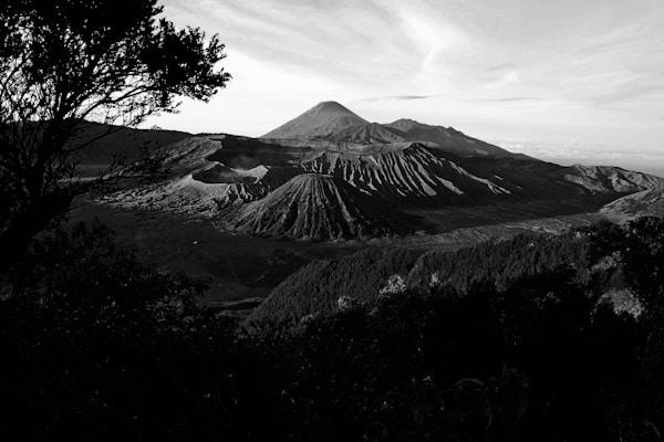 A dramatic black and white landscape photograph featuring a series of rugged volcanic mountains under a vast, partly cloudy sky. The foreground is dominated by silhouetted trees, adding depth to the composition. The central mountain stands prominently with eroded ridges and valleys.