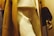 Close-up of a mannequin dressed in a beige coat with dark brown fur cuffs, cream-colored pants, and a light-colored shirt. The mannequin's hand is visible on the right side of the image.