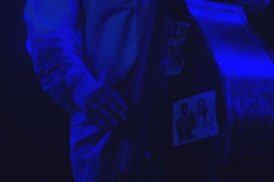 A close-up of a musician's hand playing a double bass, adorned with black-and-white photographs of jazz musicians. The scene is illuminated with a blue light, creating a moody and atmospheric effect.