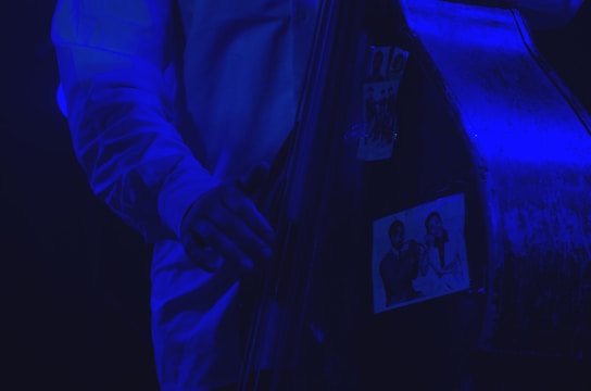 A close-up of a musician's hand playing a double bass, adorned with black-and-white photographs of jazz musicians. The scene is illuminated with a blue light, creating a moody and atmospheric effect.