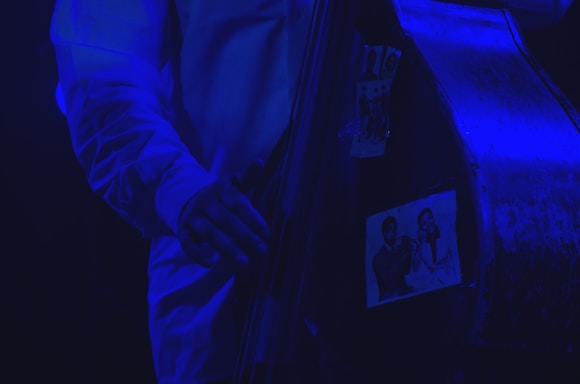 A close-up of a musician's hand playing a double bass, adorned with black-and-white photographs of jazz musicians. The scene is illuminated with a blue light, creating a moody and atmospheric effect.