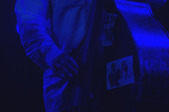 A close-up of a musician's hand playing a double bass, adorned with black-and-white photographs of jazz musicians. The scene is illuminated with a blue light, creating a moody and atmospheric effect.