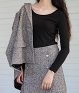 A person is wearing a black long-sleeve top paired with a gray, textured skirt and draped fabric over one shoulder. The outfit features decorative buttons, and the overall pattern has a tweed-like appearance. The setting is plain with a light-colored background.