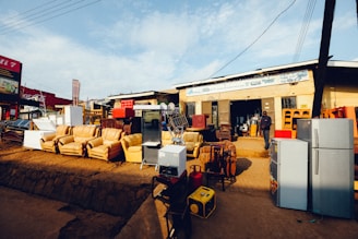 A storefront features various household appliances and furniture. Several refrigerators are placed outside, alongside couches and chairs. The background shows a building with signage and open doorways. A person stands near the entrance.