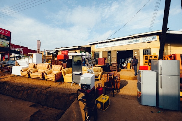 A storefront features various household appliances and furniture. Several refrigerators are placed outside, alongside couches and chairs. The background shows a building with signage and open doorways. A person stands near the entrance.
