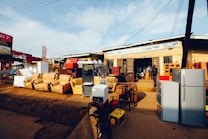 A storefront features various household appliances and furniture. Several refrigerators are placed outside, alongside couches and chairs. The background shows a building with signage and open doorways. A person stands near the entrance.