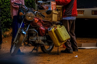 A motorcycle is parked on a brick surface, equipped with yellow jerry cans hanging on the side and a wooden crate filled with boxes on the back. Two people are interacting near the bike, one partially visible from the waist down, wearing a red jacket. The scene includes a parked car in the background and some greenery on the left side.