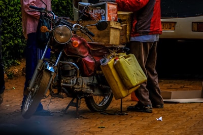 A motorcycle is parked on a brick surface, equipped with yellow jerry cans hanging on the side and a wooden crate filled with boxes on the back. Two people are interacting near the bike, one partially visible from the waist down, wearing a red jacket. The scene includes a parked car in the background and some greenery on the left side.