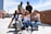 A group of five people pose energetically on a rooftop with a cityscape in the background. The people at the front are in crouched positions, displaying playful hand gestures, while two stand at the back smiling. The urban environment features a tall tower and various buildings under a partly cloudy sky.