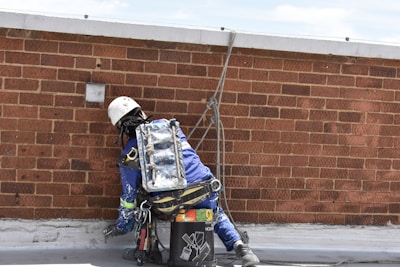 A worker in protective gear and a helmet is engaged in maintenance or construction work on a flat rooftop, using a rope for safety. The worker wears a blue uniform and is surrounded by tools and materials for the task.