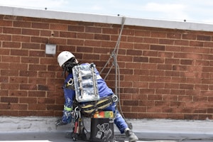 A worker in protective gear and a helmet is engaged in maintenance or construction work on a flat rooftop, using a rope for safety. The worker wears a blue uniform and is surrounded by tools and materials for the task.