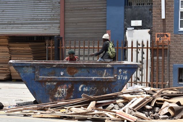 Two individuals are near a large, rusty blue dumpster placed in an industrial or urban setting. One person is sitting inside the dumpster, while the other, wearing a white beanie and carrying a backpack, leans on the edge. A stack of wooden planks and debris is scattered in the foreground. The backdrop includes corrugated metal doors, a brick wall, and a sign indicating security services.