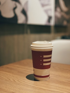 A disposable coffee cup with a red and white design is placed on a wooden table. The background is softly blurred, featuring abstract art in black and white tones, giving a cozy cafe atmosphere.