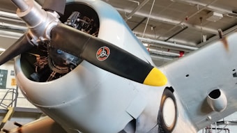 Close-up of an aircraft's propeller and engine with a yellow-tipped black blade. Visible details include mechanical components, part of the fuselage, and a hangar setting with overhead pipes and lights.