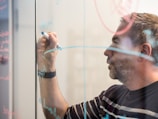 A person writing on a glass board with a blue marker. The board is covered with various writings and diagrams, possibly related to brainstorming or planning.