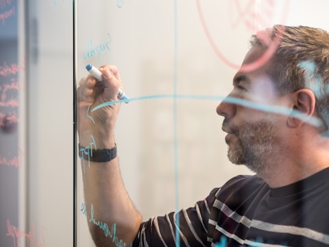A person writing on a glass board with a blue marker. The board is covered with various writings and diagrams, possibly related to brainstorming or planning.