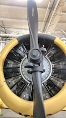 A close-up view of an aircraft engine with a prominent propeller at the center. The engine's circular housing has a yellow outer rim, contrasting with the central metallic and black components. The intricate design of the engine's internal parts, including several visible cylinders and mechanical elements, suggests it's a radial engine typical of older aircraft.