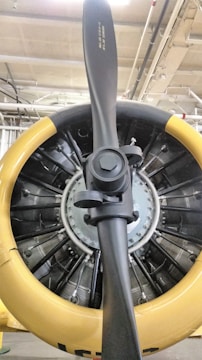 A close-up view of an aircraft engine with a prominent propeller at the center. The engine's circular housing has a yellow outer rim, contrasting with the central metallic and black components. The intricate design of the engine's internal parts, including several visible cylinders and mechanical elements, suggests it's a radial engine typical of older aircraft.